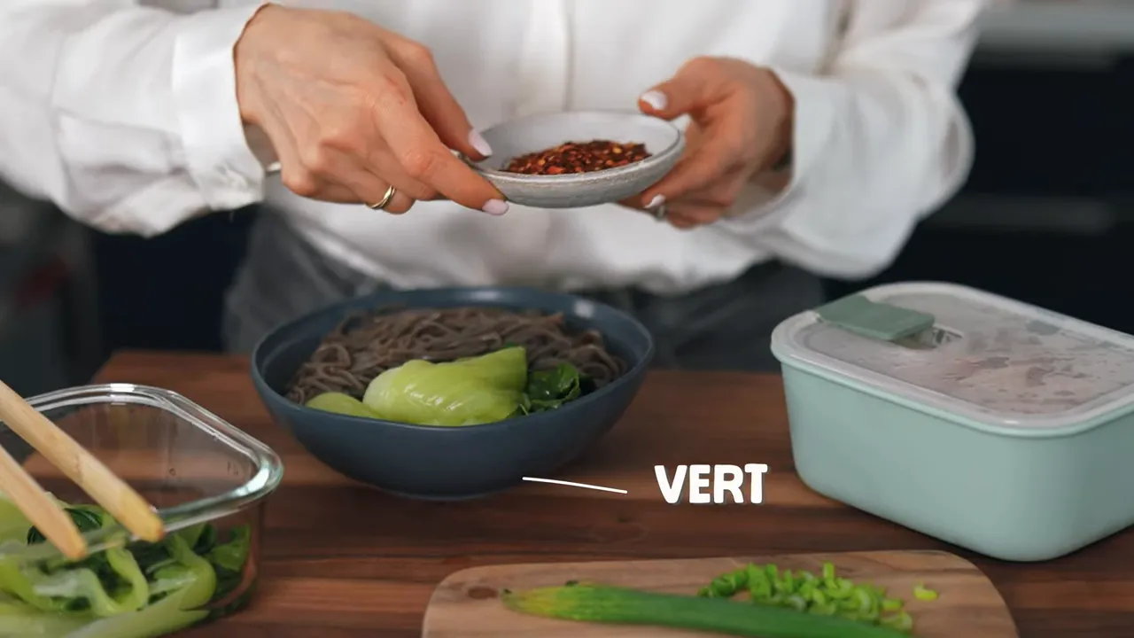 Bol de nouilles soba avec légumes verts, mains ajoutant des flocons de piment au-dessus